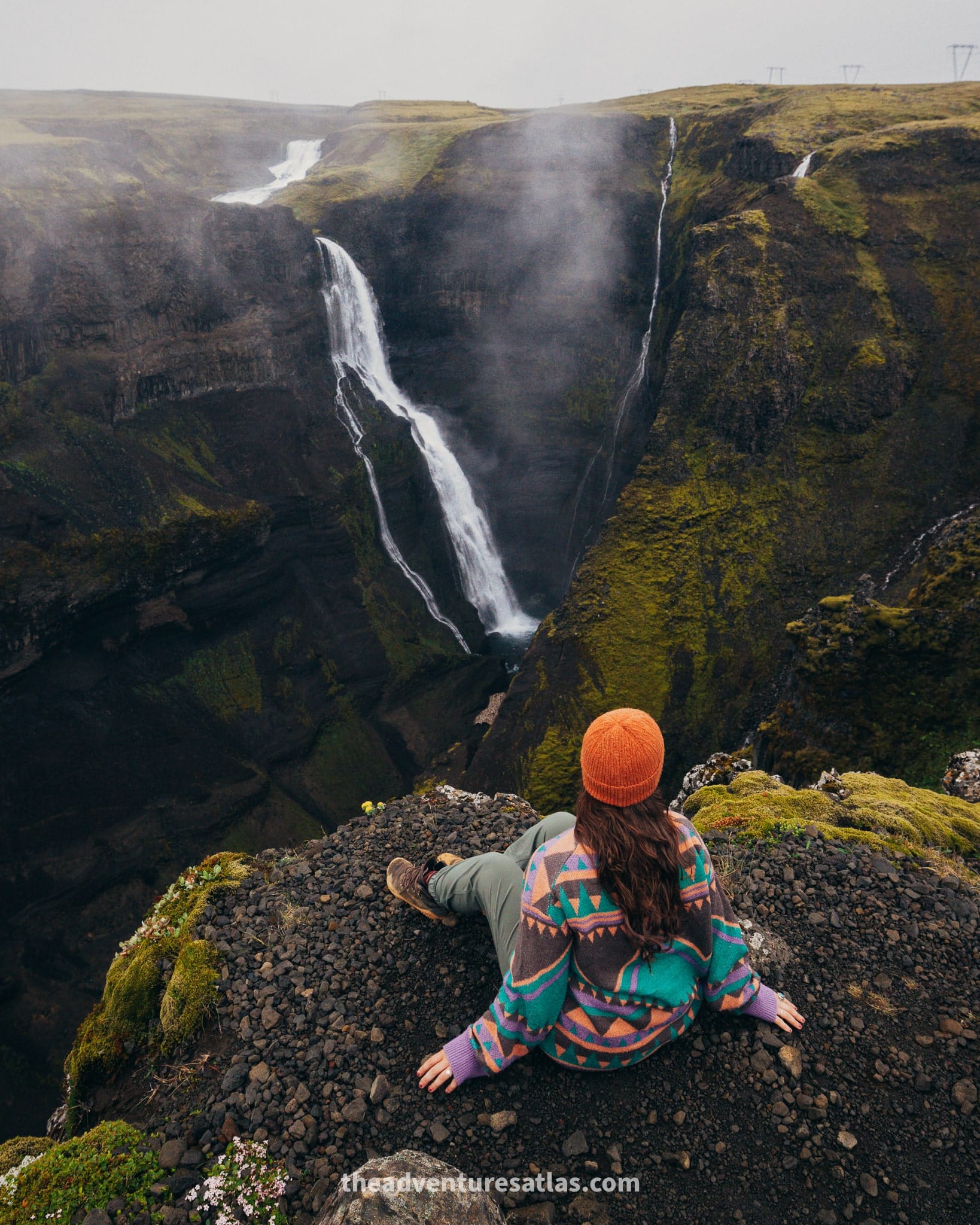 A girl sitting on the edge of a waterfall in Iceland wearing a sweater, beanie, and waterproof hiking boots