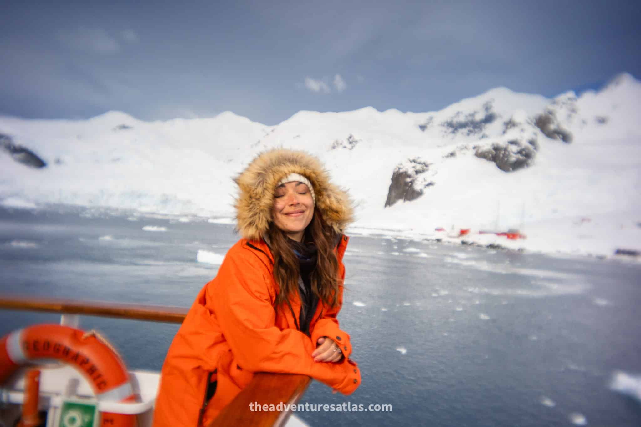 Traveler in an orange expedition parka leaning on a ship railing with Antarctic mountains and floating ice in the background.