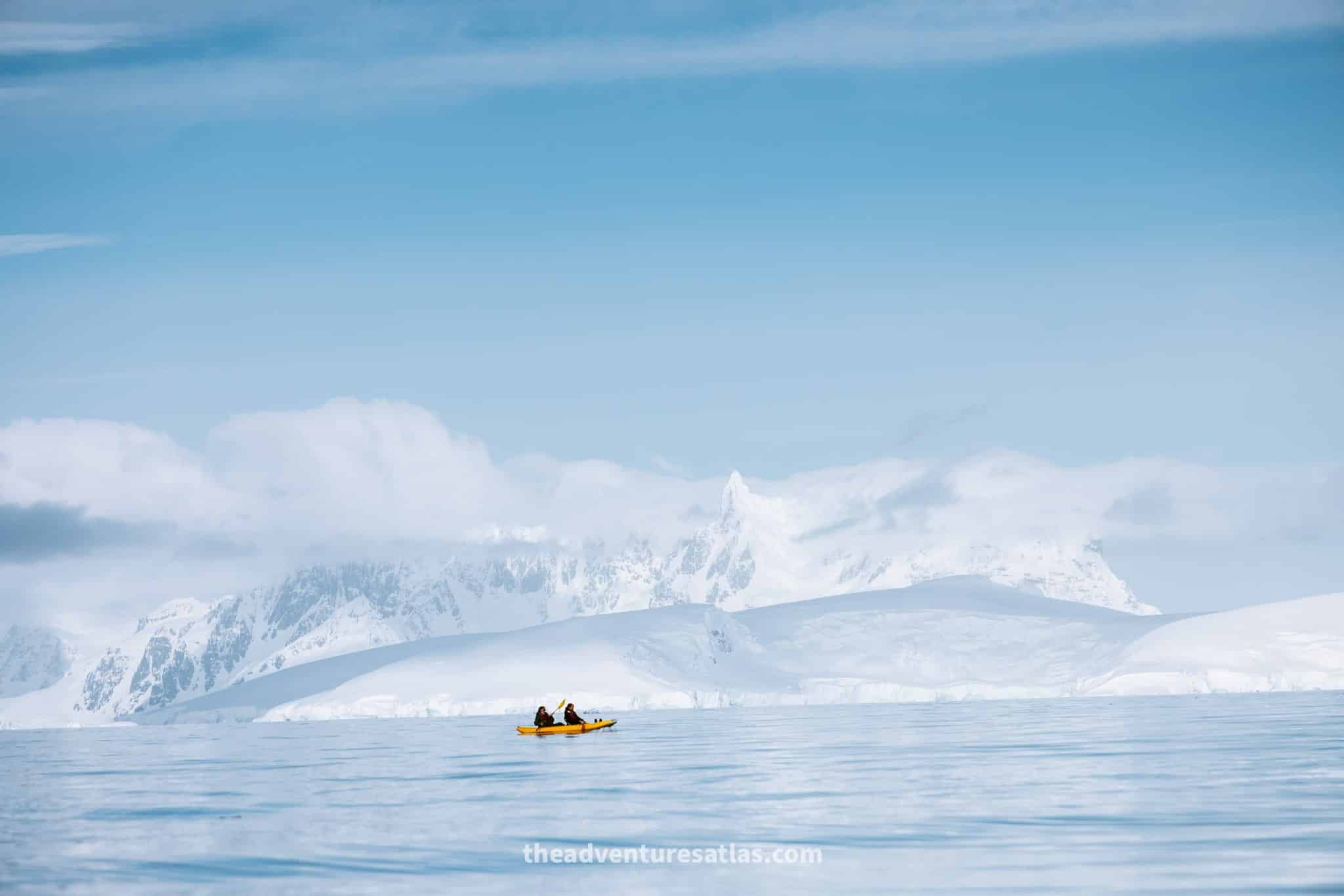 Two passengers kayaking in a yellow kayak on a calm sea in Antarctica with tall snow covered mountains in the background