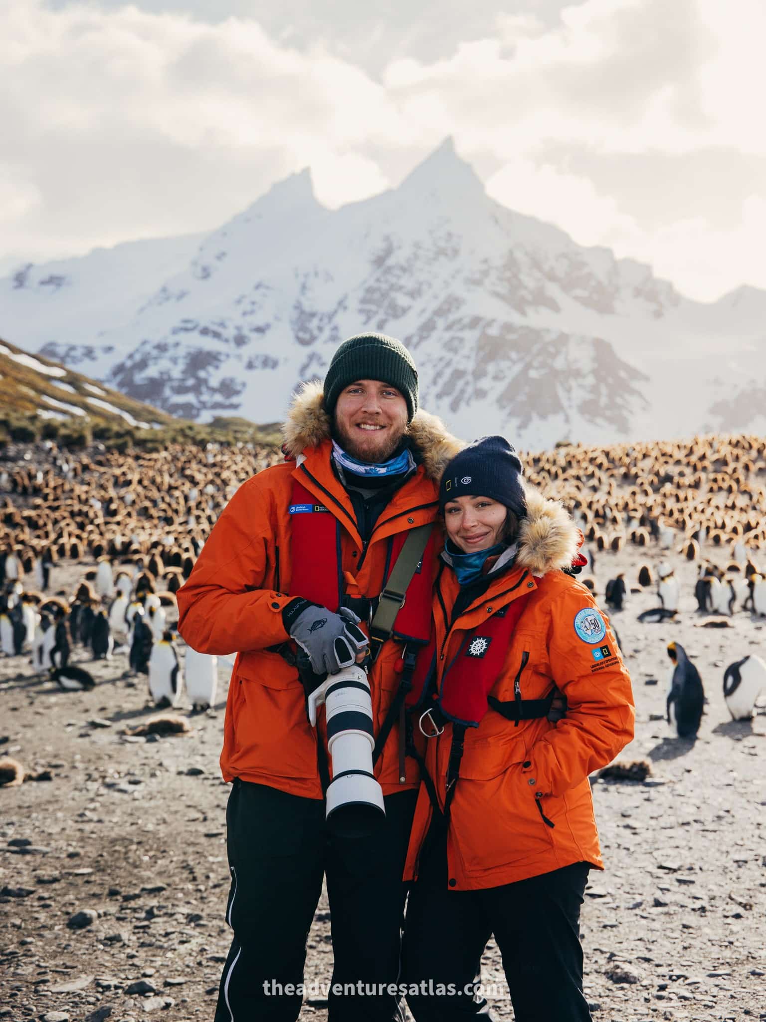 A couple in orange National Geographic-Lindblad parkas holding cameras in front of thousands of penguins at sunset on South Georgia Island