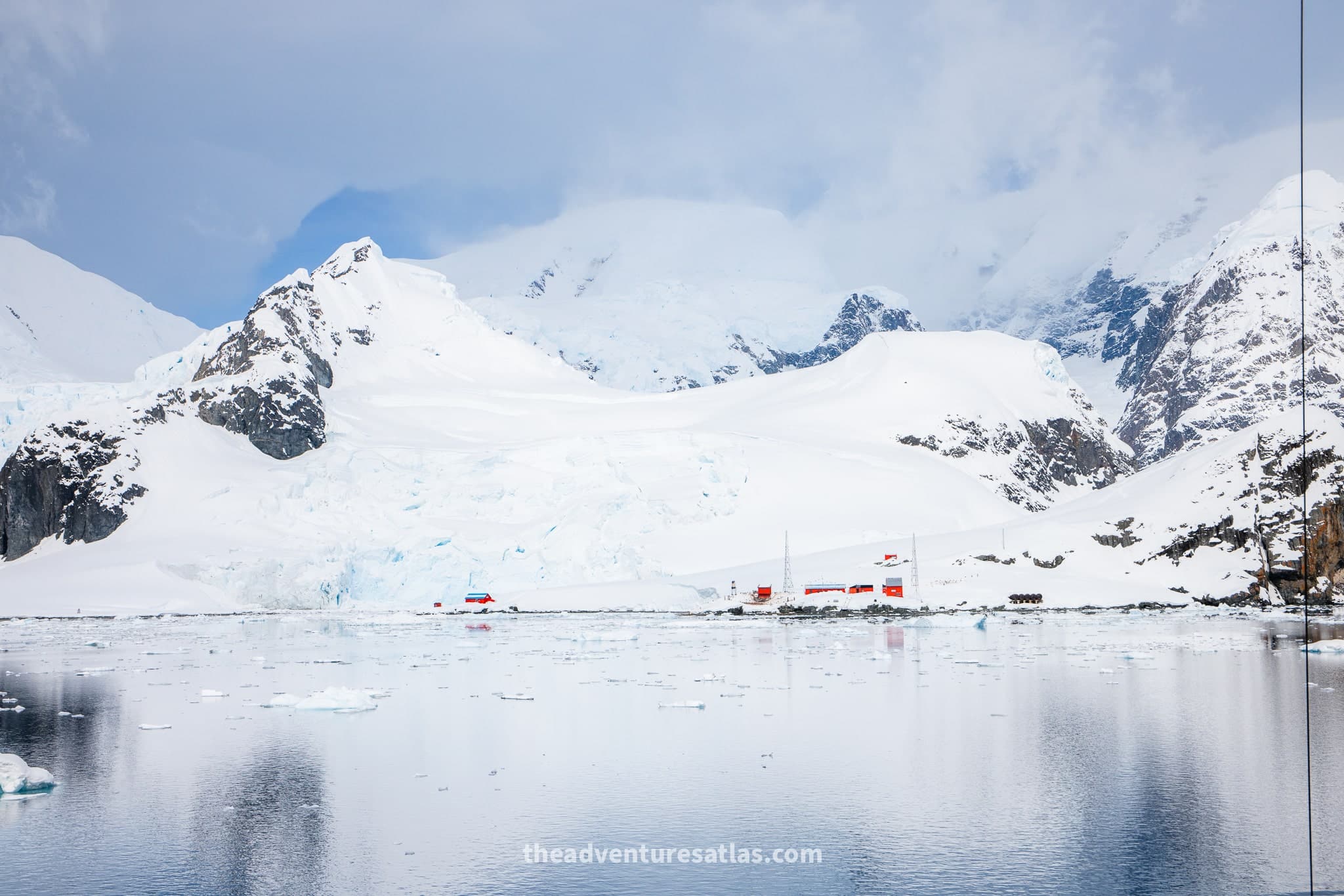 Antarctic research station along the shoreline with snow-covered mountains and ice-filled water in the foreground.